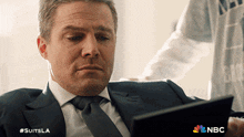 a man in a suit and tie is looking at a tablet with the nbc logo on the bottom right