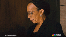 a woman wearing glasses stands in front of a wooden wall with the nbc logo on the bottom