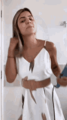 a woman in a white dress is blow drying her hair while standing in front of a mirror .