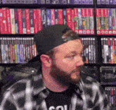 a man with a beard is wearing a baseball cap and a plaid shirt in front of a bookshelf .