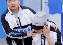 a man wearing an adidas jacket is petting another man 's face