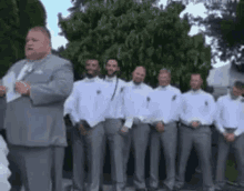a man in a suit and tie is standing next to a group of men in white shirts and gray pants .