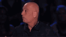 a bald man is sitting in a dark room with his mouth open and a crowd behind him .