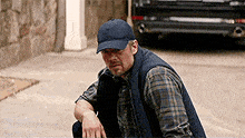 a man wearing a plaid shirt and a baseball cap is kneeling down on the sidewalk .