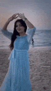 a woman in a blue dress stands on a beach with her arms in the air
