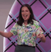 a woman in a floral shirt is standing in front of a purple wall and making a funny face .