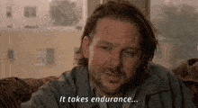 a man with long hair and a beard is sitting on a couch and saying it takes endurance