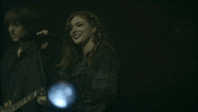 a woman singing into a microphone with a man playing a guitar behind her