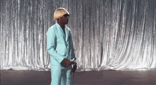 a man in a light blue suit and sunglasses is standing in front of a silver curtain .