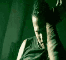 a man in a black tank top is sitting in a dark room with his hands on his head .