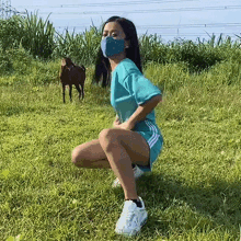 a woman wearing a mask squatting in a field with a horse in the background .