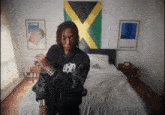a man standing in front of a bed with a jamaican flag hanging on the wall behind him