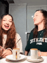 two girls are sitting at a table with two cups of coffee and one girl is wearing a shirt that says athletic