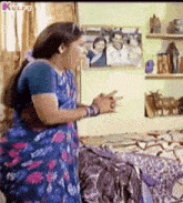 a woman in a blue saree is standing next to a bed holding a cell phone in her hand .