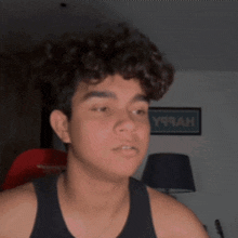 a young man with curly hair looks at the camera with a sign in the background that says y99ah