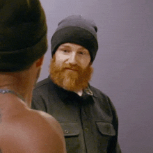 a man with a beard is wearing a beanie and looking at another man .