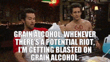 two men are sitting at a table with bottles of alcohol . one of the men has the word go painted on his chest .