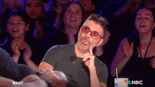 a man wearing glasses sits in a crowd of people with a nbc cup in the foreground