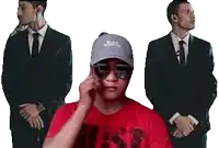 a man wearing sunglasses and a hat is talking on a cell phone while two men in suits stand behind him .