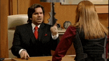 a man in a suit and tie is pointing at a woman in a red shirt while sitting at a desk .