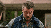 a man with a beard and a plaid shirt is wearing a vest and looking down .