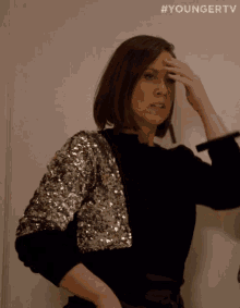 a woman wearing a black top and a silver sequined jacket holds her hand to her forehead .
