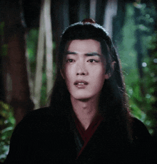 a young man with long black hair is standing in a forest looking at the camera .