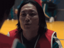 a man with long hair is wearing a red vest and talking to another man .