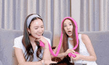 two women sitting on a couch one wearing a headband and the other holding a pink braided object