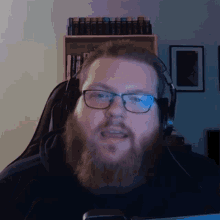 a man with a beard wearing glasses and headphones is sitting in front of a computer screen .