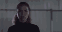 a woman in a black turtleneck is standing in a dark room looking at the camera .