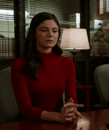 a woman in a red turtleneck sits at a table with her hands folded
