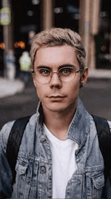 a young man wearing glasses and a denim jacket is standing in front of a building .
