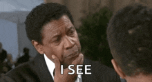 a man in a suit and tie is talking to another man while holding his hand to his chin .
