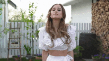 a woman in a white top and skirt stands in front of a white fence