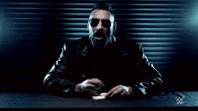 a man in a leather jacket sits at a table with a w logo in the background