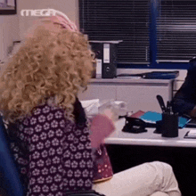 a woman with curly blonde hair is sitting at a desk talking on a cell phone .