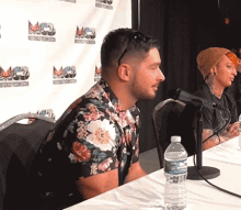 a man in a floral shirt sits at a table with a bottle of water in front of him