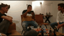 a group of men are sitting around a table with bottles of beer and snacks and one man is covering his face with his hand