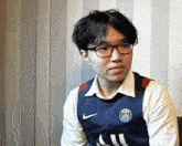 a young man wearing glasses and a blue nike jersey is sitting in front of a wall .