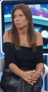 a woman in a black off the shoulder top and leather pants is sitting in a chair with her hands folded .