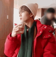 a young man wearing a red jacket and a white beanie is drinking a cup of coffee .