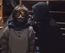a person wearing a mask and goggles stands next to another person wearing a hoodie