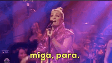 a woman in a pink jacket is singing into a microphone with the words miga para on the bottom .
