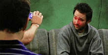 a man is sitting on a couch taking a picture of another man with a cell phone .