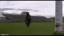 a man in a trench coat is walking on a field next to a cigarette butt .