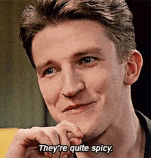 a man is smiling and saying `` they 're quite spicy '' while sitting on a couch .