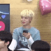 a man with blonde hair is holding a cell phone in his hand while sitting in a room with other people .