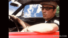 a man wearing sunglasses and a hat is driving a red car and the website gifrun.com is below him