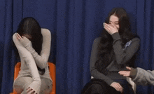 two women are sitting next to each other in front of a blue curtain . one of the women is covering her face with her hand .